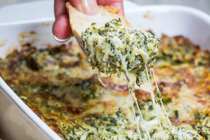 A close up of a piece of bread coming out of the dip with a string of cheese stretching from the dish