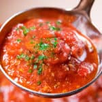 A close-up of a ladle filled with vibrant tomato sauce, garnished with fresh parsley. The chunky texture highlights pieces of tomato. In the background, a blur of rich red sauce simmers in the pot.