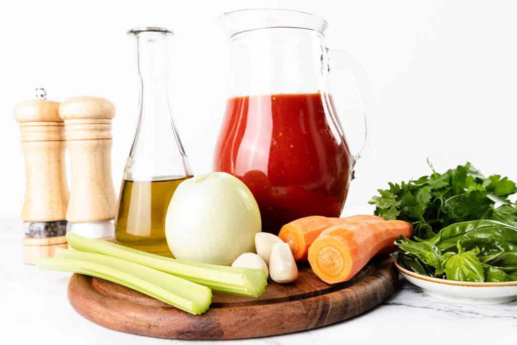 A wooden board holds a whole onion, two carrots, two celery stalks, three garlic cloves, a small bottle of olive oil, a pitcher of tomato sauce, and a plate of fresh herbs, with salt and pepper shakers behind—perfect for homemade tomato sauce.