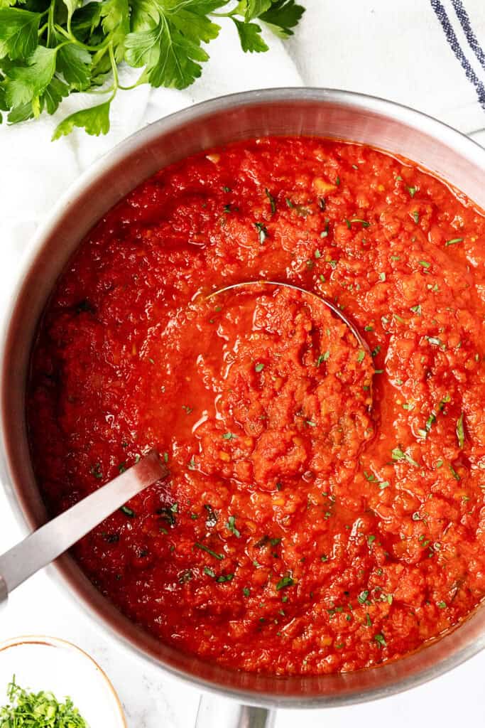 A pot filled with chunky red tomato sauce, garnished with fresh herbs, with a ladle resting inside. Fresh parsley and a small bowl of chopped herbs sit nearby on a white surface, enhancing the classic appeal of homemade tomato sauce.