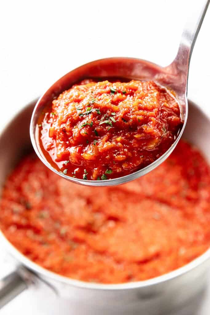 A close-up of a ladle lifting chunky red tomato sauce, topped with herbs, over a saucepan brimming with rich tomato sauce.