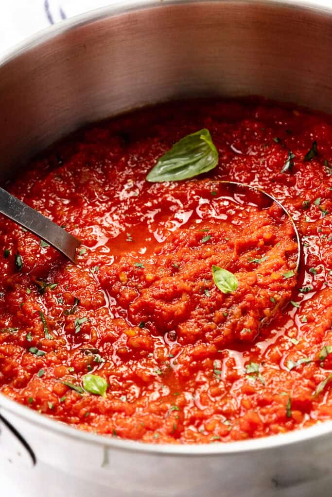 A close-up of a pot filled with chunky Tomato Sauce, garnished with fresh basil leaves. A ladle rests in the rich, red sauce, highlighting its vibrant color and homemade texture.