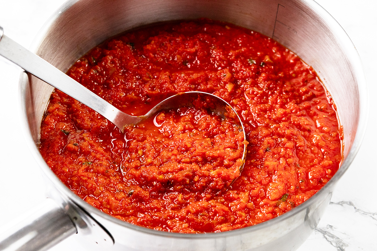 A stainless steel saucepan filled with chunky Tomato Sauce, thick and vibrant, with visible herbs and oil glistening on the surface. A metal ladle rests inside, ready to serve this rich and flavorful creation.
