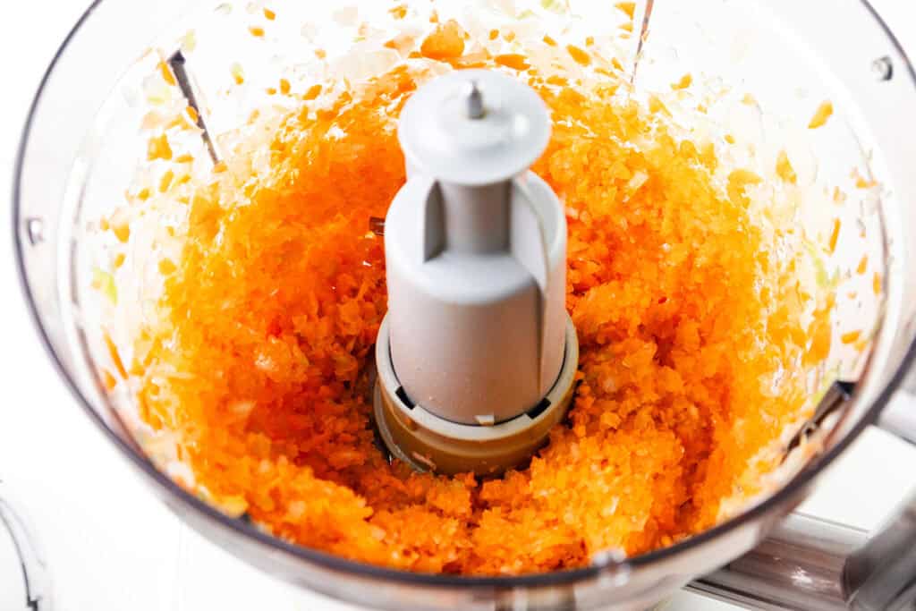 Finely chopped orange carrots and white onions inside a food processor bowl, viewed from above. The evenly processed vegetables surround the central blade—perfectly prepped for adding to a homemade tomato sauce.
