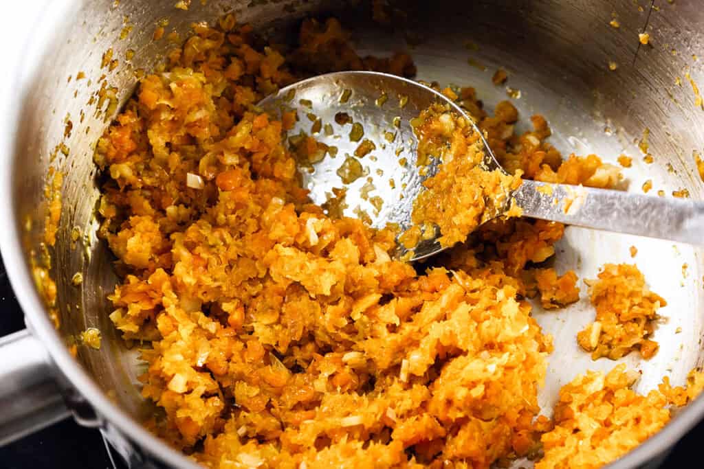 Chopped carrots, onions, and celery sauté in a pot, being stirred with a metal spoon. The vegetables are softened and lightly browned before rich tomato sauce is added, forming the base for a savory dish.