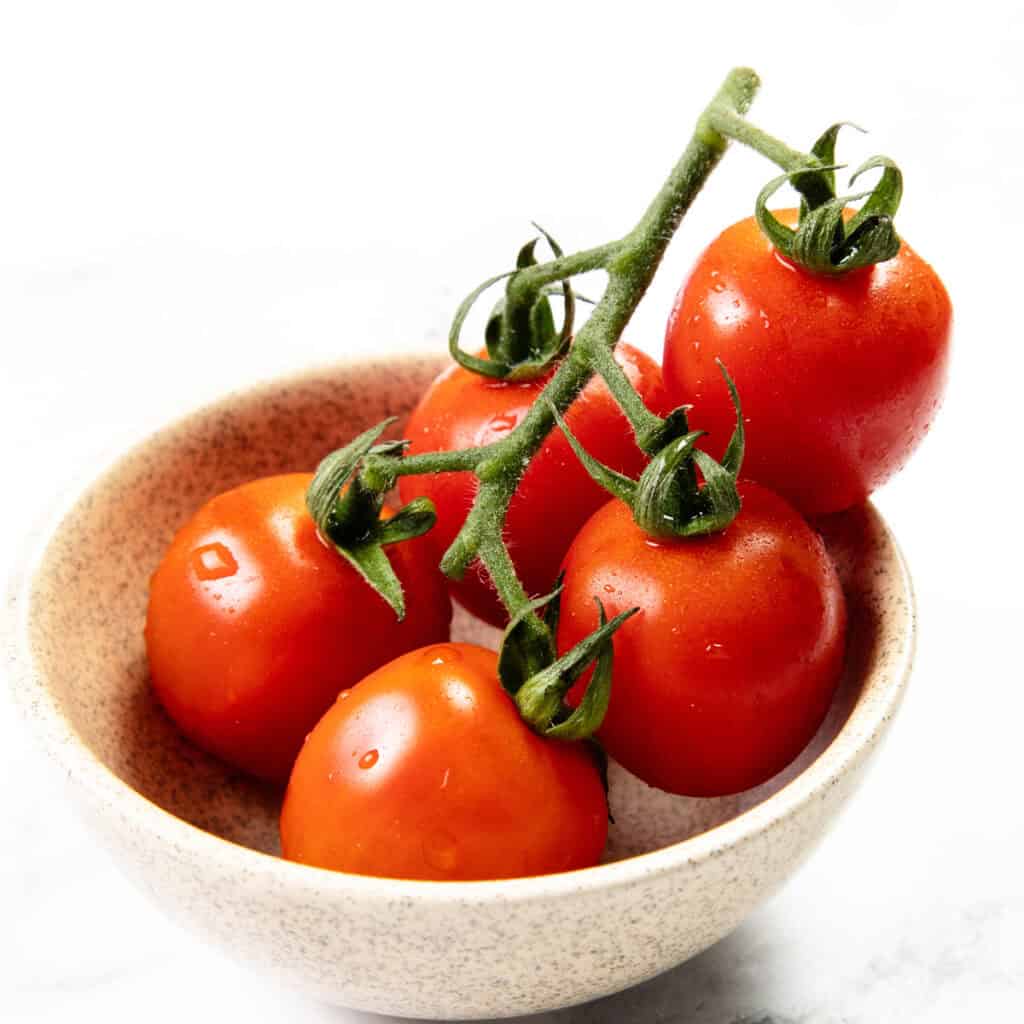 A small beige bowl holds five fresh red tomatoes still attached to their green vine, perfect for making homemade tomato sauce, set against a white background.