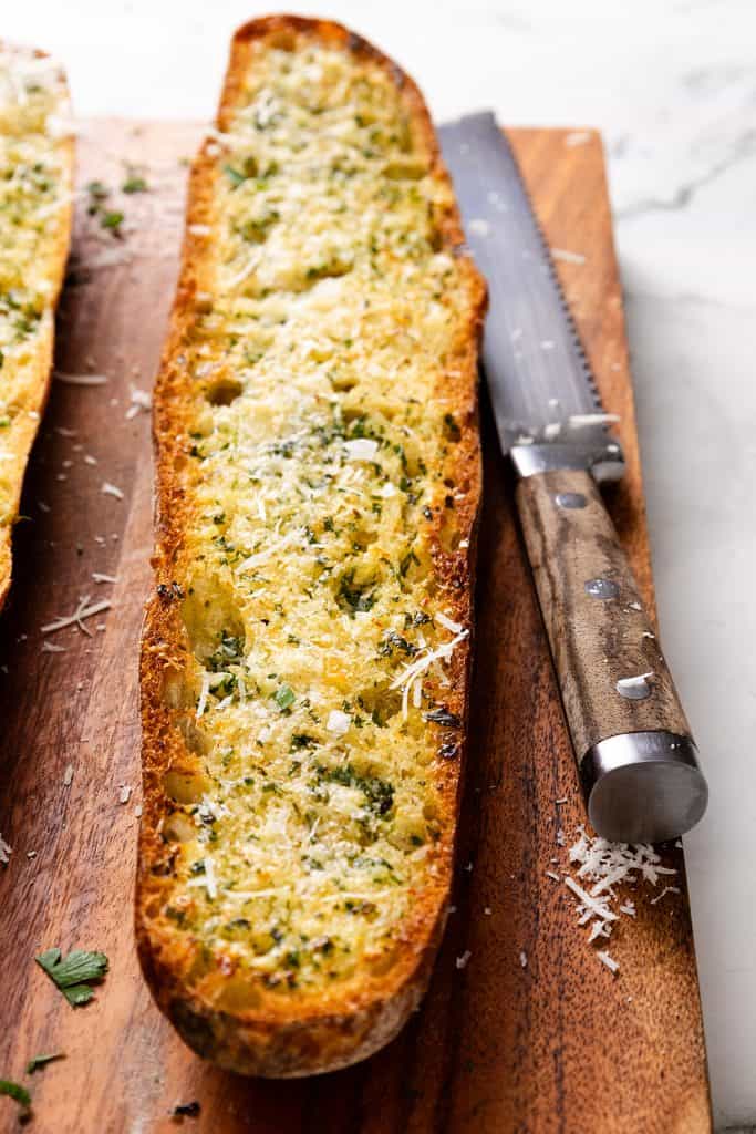 A sliced loaf of perfect homemade garlic bread topped with herbs and grated cheese sits on a wooden cutting board beside a bread knife.
