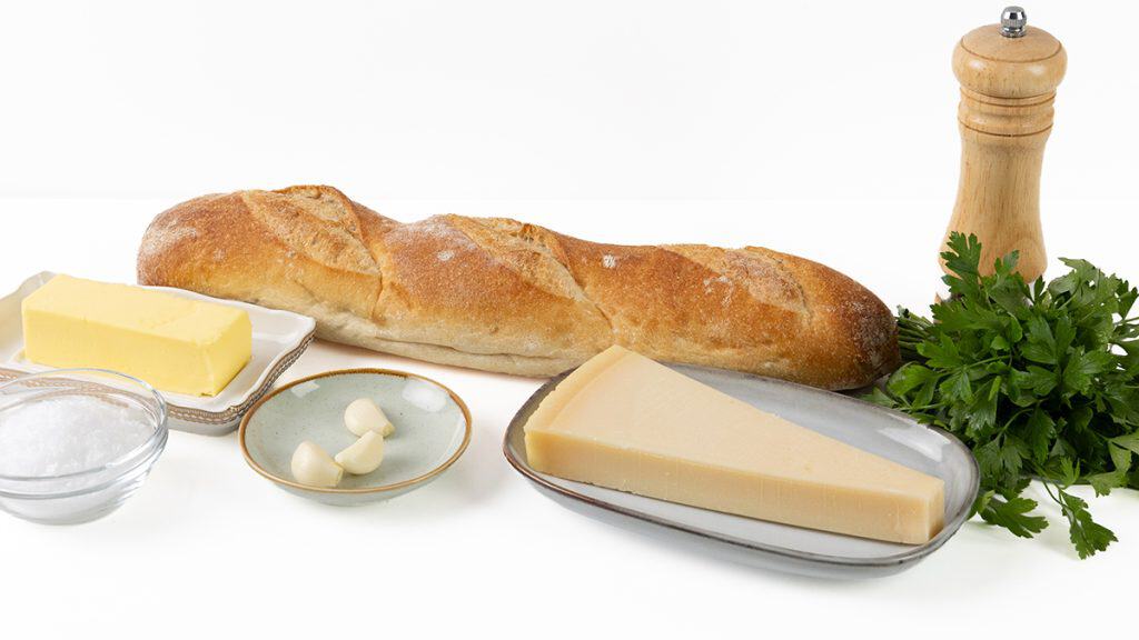A baguette, a wedge of cheese, butter, garlic cloves, a bowl of salt, fresh parsley, and a wooden pepper grinder are arranged on a white surface&mdash;just the ingredients you need for perfect homemade garlic bread.