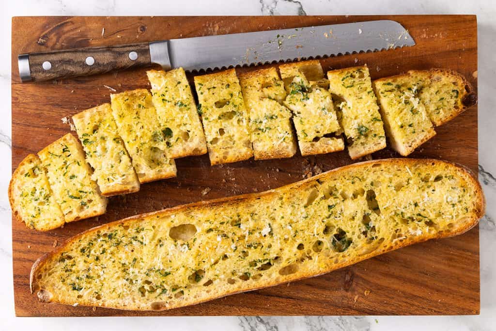 Perfect Homemade Garlic Bread awaits: sliced with herbs and grated cheese on a wooden cutting board, next to a serrated knife. One half is cut into pieces, the other remains whole&mdash;delivering irresistible flavor in every bite.
