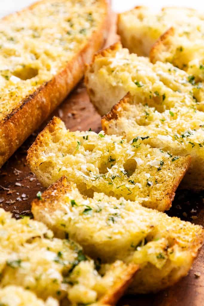 Slices of golden, crispy Perfect Homemade Garlic Bread topped with herbs and grated cheese are arranged on a wooden board.