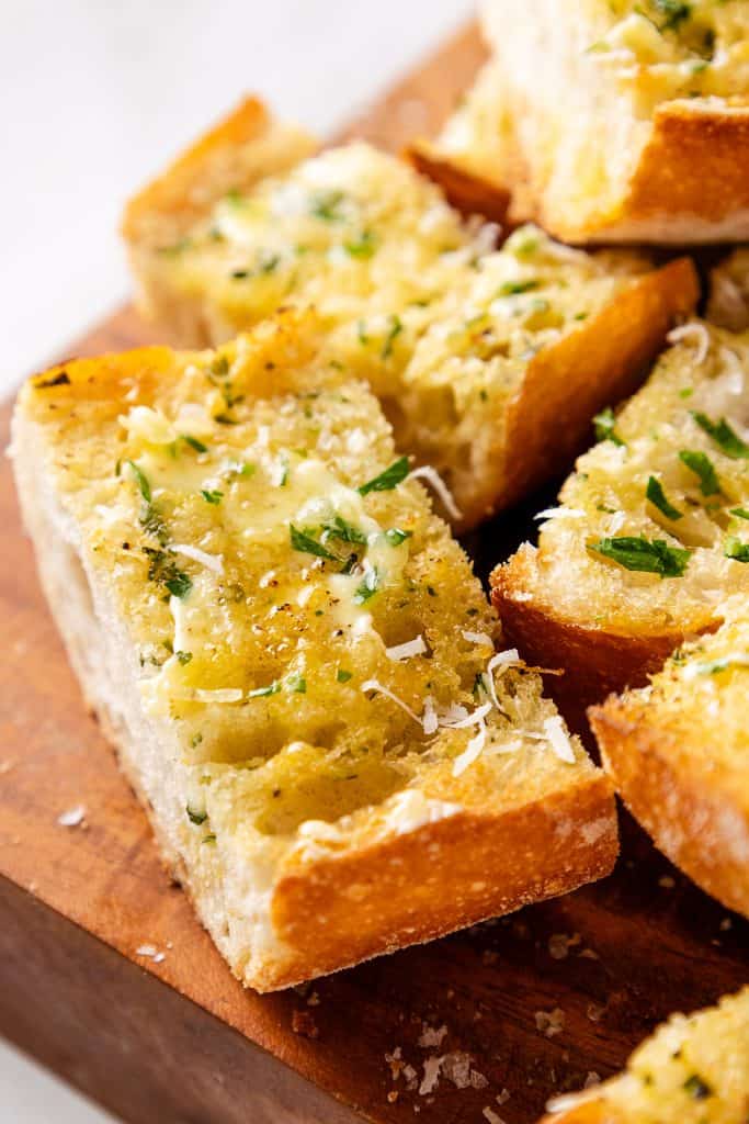 Close-up of Perfect Homemade Garlic Bread, featuring sliced bread with melted butter, chopped parsley, and grated cheese on top, served on a wooden board.