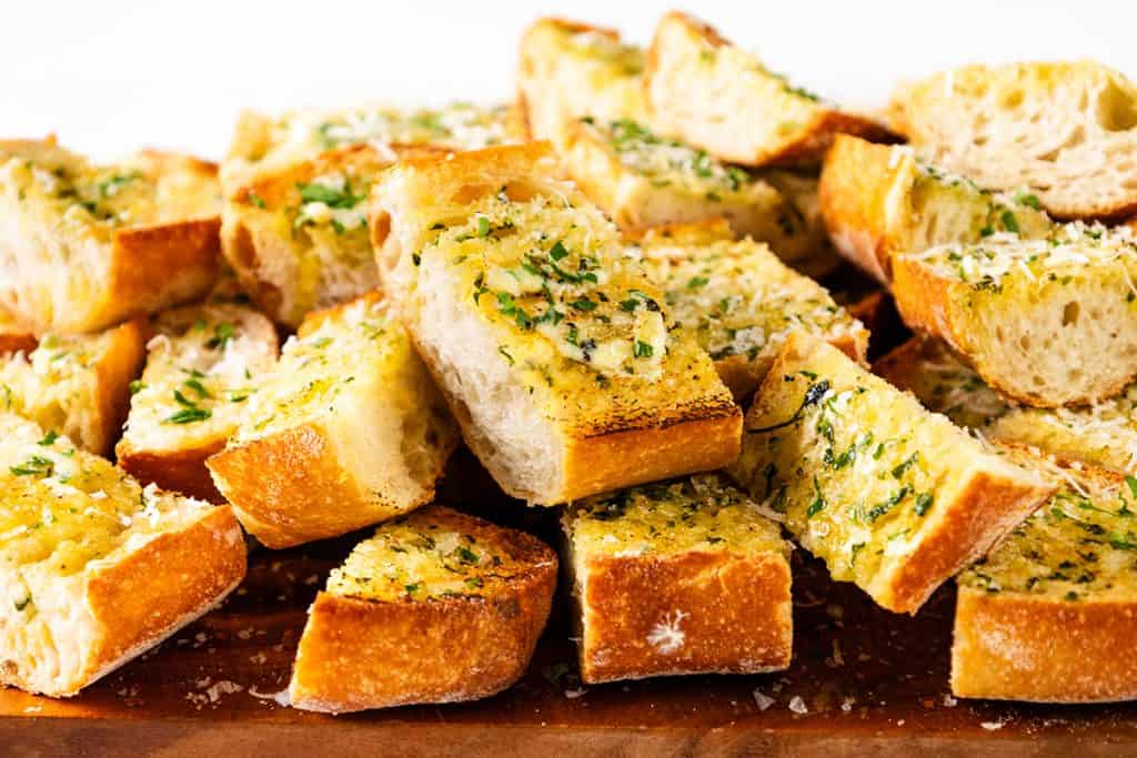 Sliced pieces of golden, crispy Perfect Homemade Garlic Bread topped with chopped parsley and grated cheese, stacked on a wooden surface.