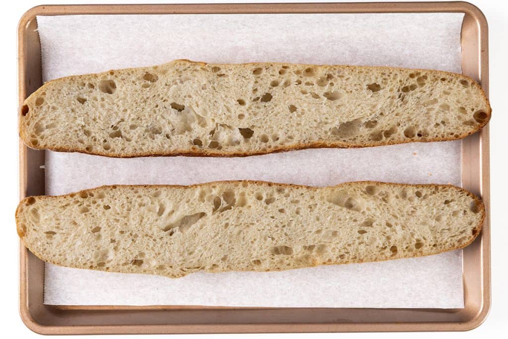 Two halves of a sliced loaf of bread are placed cut-side up on a parchment-lined baking sheet, showing an airy crumb texture that&rsquo;s ideal for creating the Perfect Homemade Garlic Bread.
