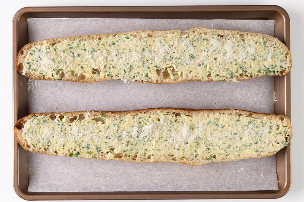 A baking tray with two halves of a baguette, topped with a garlic herb butter mixture and grated cheese, is ready to be baked into the perfect homemade garlic bread. The tray is lined with parchment paper.