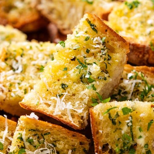 Perfect Homemade Garlic Bread slices, golden-brown and topped with chopped herbs and grated cheese, are stacked closely together. The bread looks crispy on the outside and soft on the inside.