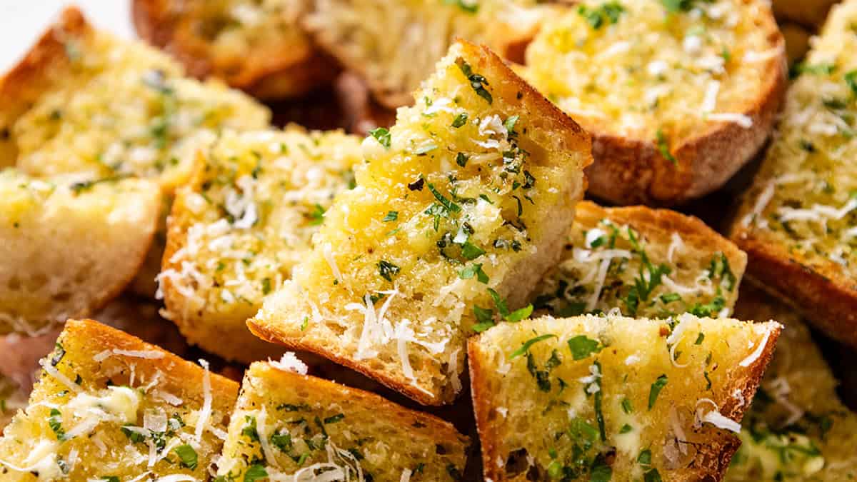 Perfect Homemade Garlic Bread slices, golden-brown and topped with chopped herbs and grated cheese, are stacked closely together. The bread looks crispy on the outside and soft on the inside.