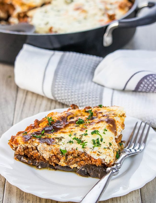 A slice of Moussaka on a white plate with the pan in the background