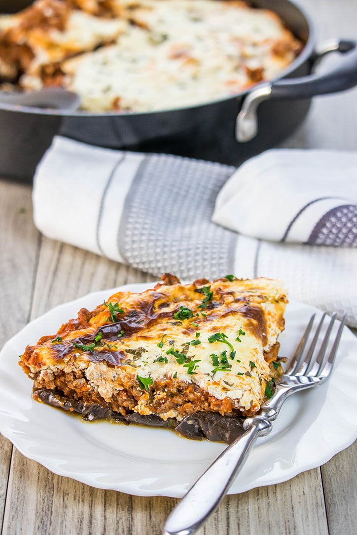 A slice of Moussaka on a white plate with the pan in the background