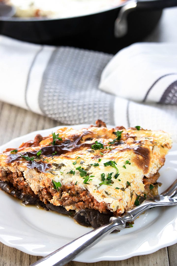 A slice of Moussaka on a white plate, and a fork next to it
