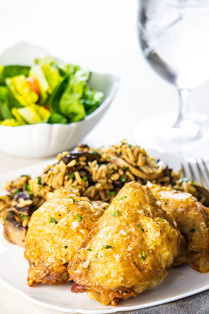 cooked chicken thighs on a white plate with rice and salad in the background