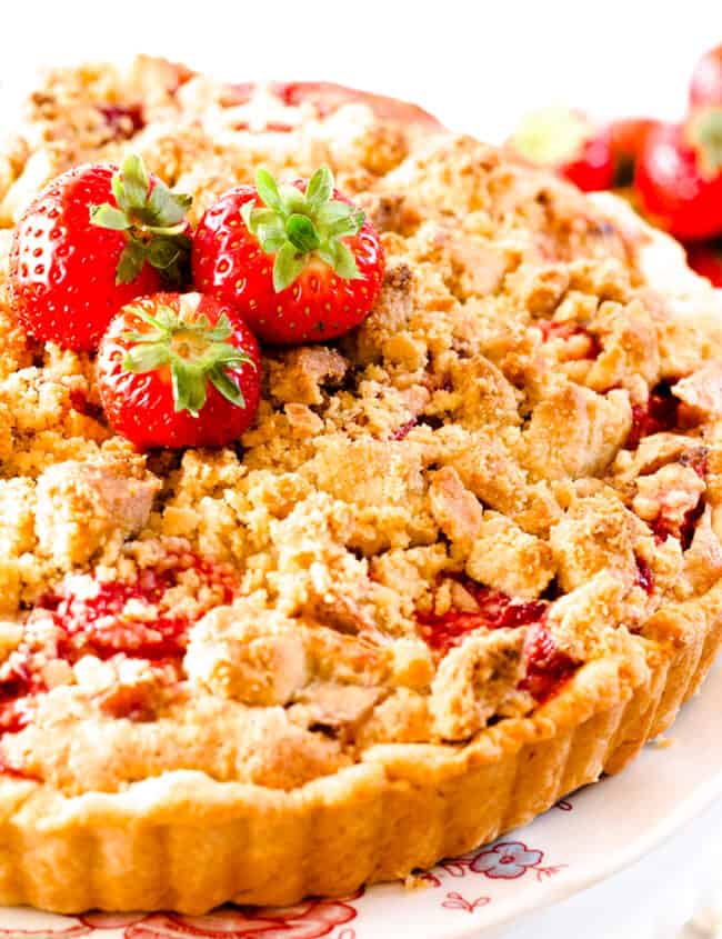 A close up of a Strawberry Rhubarb Pie with a crumb topping on a serving plate topped with strawberries.