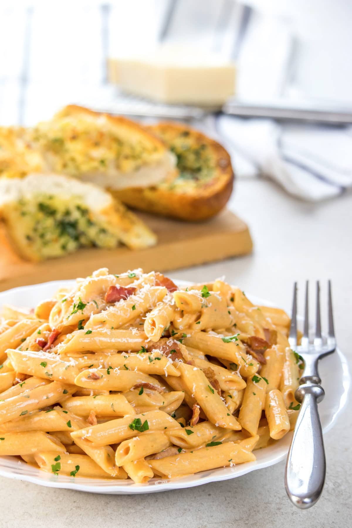 Penne Alla Vodka piled high on a white plate, sprinkled with parmasean cheese anf garlic bread in the background