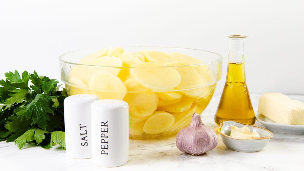A glass bowl of sliced potatoes for Crispy Garlic Roasted Potatoes, a bunch of parsley, salt and pepper shakers, a garlic bulb with cloves, a bottle of oil, and a slab of butter on a white surface.