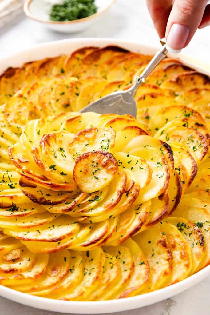 A hand lifts a serving of Crispy Garlic Roasted Potatoes—thinly sliced, golden-brown, and arranged in a circular pattern in a white dish, garnished with chopped herbs.
