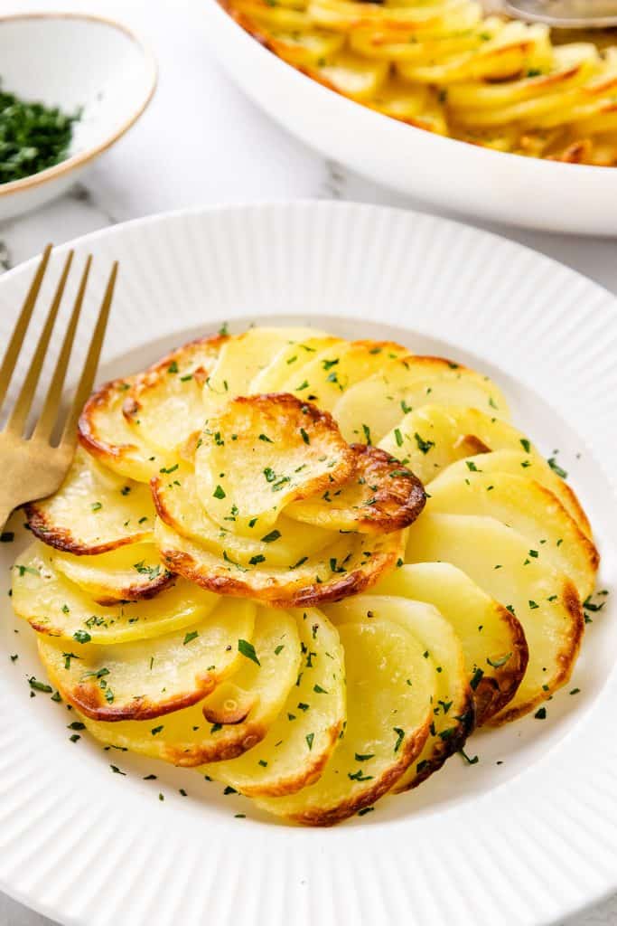 A white plate with thinly sliced, golden-brown Crispy Garlic Roasted Potatoes arranged in overlapping layers, garnished with chopped parsley. A gold fork rests on the plate, and more potatoes are visible in a dish in the background.