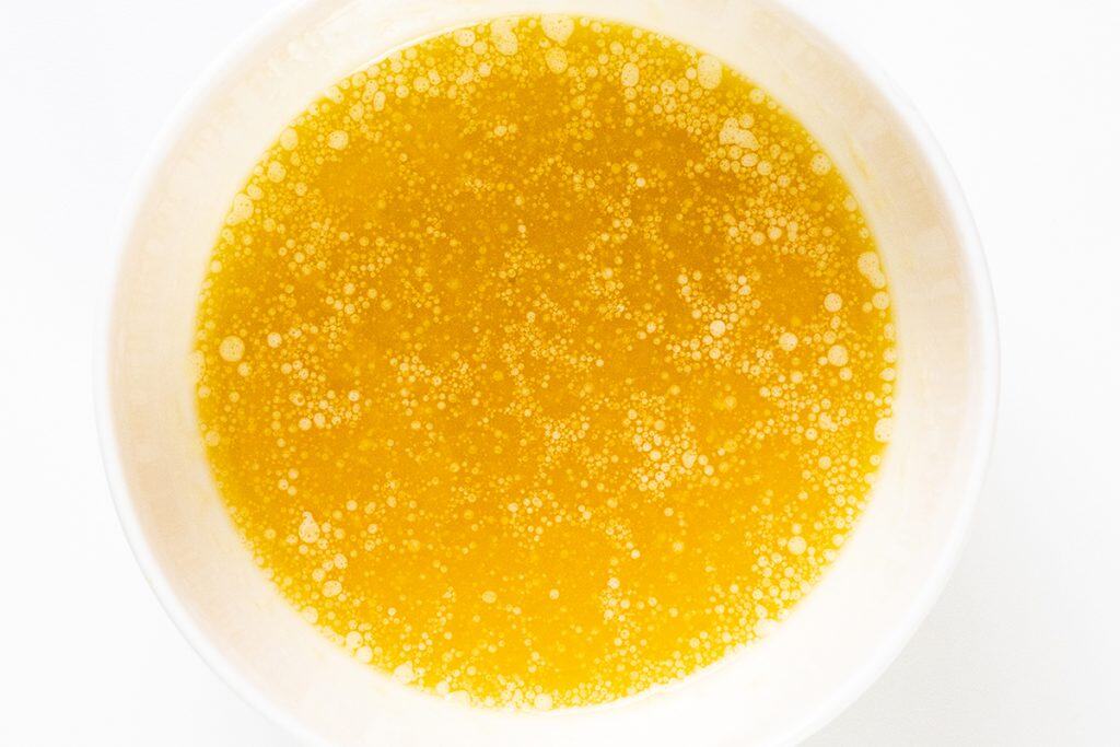 A white bowl filled with melted yellow butter, showing a layer of small bubbles and oil droplets—perfect for drizzling over Crispy Garlic Roasted Potatoes—set against a white background.