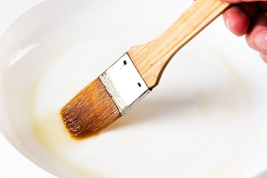 A close-up of a hand holding a paintbrush, dipped in a shallow white dish containing a translucent liquid, ready to add the finishing touch to Crispy Garlic Roasted Potatoes.