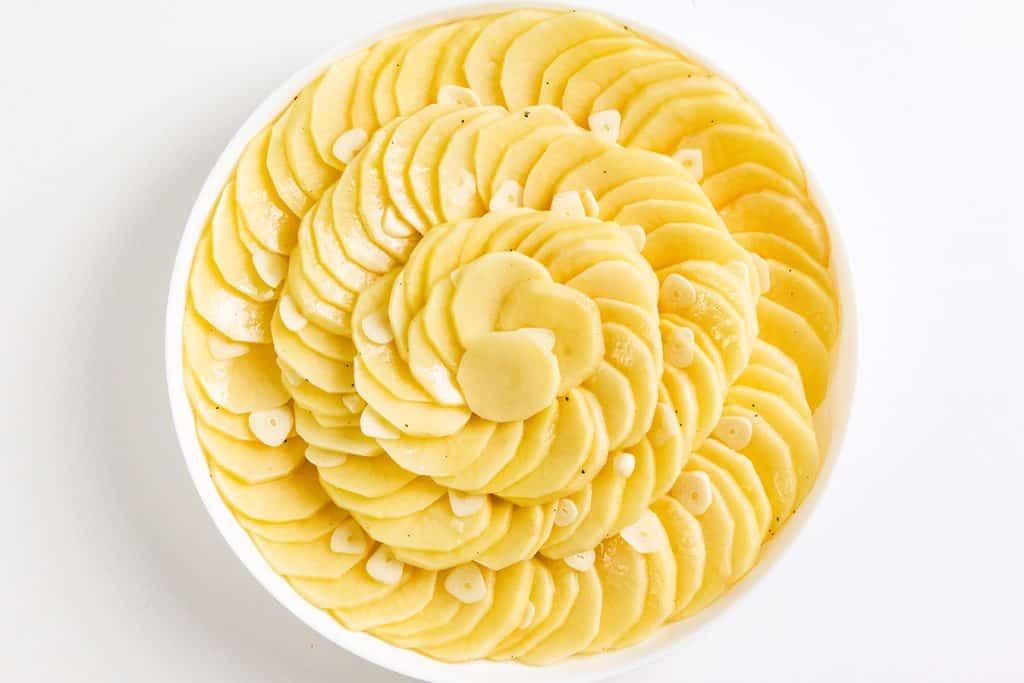 A round white dish filled with thinly sliced Crispy Garlic Roasted Potatoes, arranged in overlapping spiral layers on a white background.