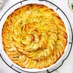 A round white dish filled with thinly sliced, golden-brown Crispy Garlic Roasted Potatoes arranged in a spiral pattern, garnished with chopped herbs. The dish sits on a white marble surface surrounded by seasonings and garlic cloves.