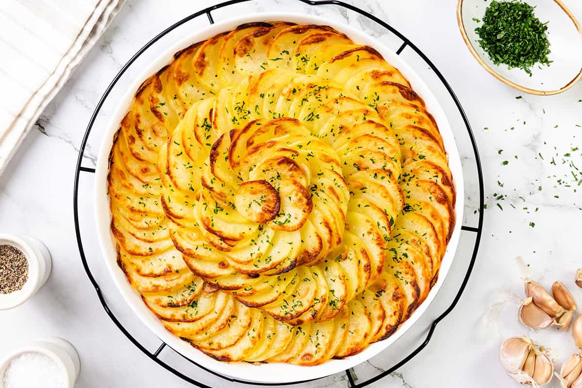 A round white dish filled with thinly sliced, golden-brown Crispy Garlic Roasted Potatoes arranged in a spiral pattern, garnished with chopped herbs. The dish sits on a white marble surface surrounded by seasonings and garlic cloves.