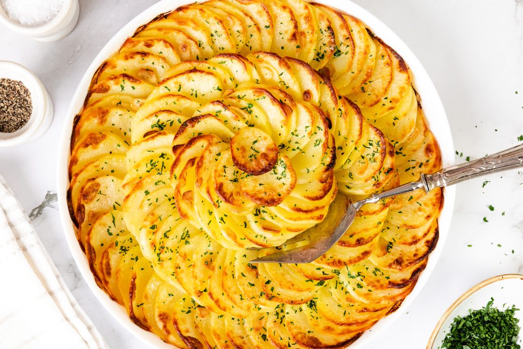 A round white dish filled with thinly sliced, golden-brown Crispy Garlic Roasted Potatoes arranged in a spiral pattern, garnished with chopped herbs, with a serving utensil resting inside.