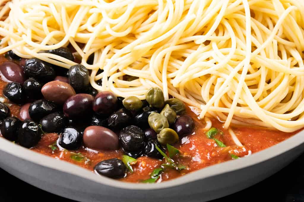 Close-up of a dish showcasing Spaghetti Alla Puttanesca with cooked spaghetti on one side and vibrant tomato sauce on the other, topped with black and kalamata olives, capers, and fresh herbs, all beautifully arranged in a white bowl.