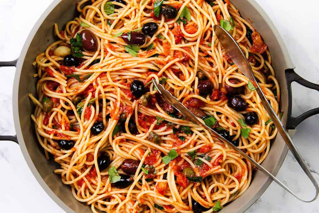 A pan filled with vibrant Spaghetti Alla Puttanesca showcases noodles mingling with rich tomato sauce, black olives, tangy capers, and a sprinkle of fresh parsley. A serving spoon and fork rest invitingly on top.