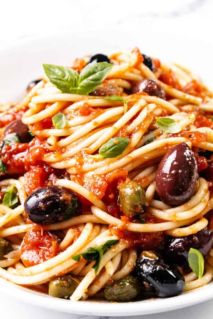 Close-up of a plate of Spaghetti Alla Puttanesca, featuring tomato sauce, olives, and capers. This vibrant dish is garnished with fresh basil leaves, adding a pop of green to the rich red sauce and the glossy black and purple olives.