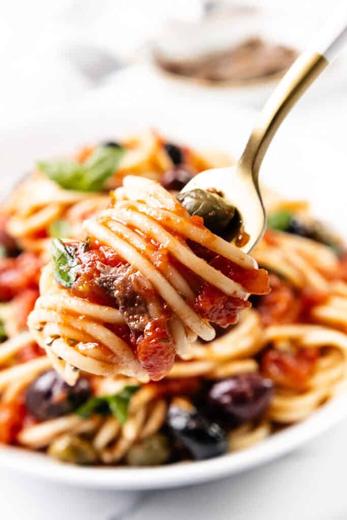 A fork holding a swirl of Spaghetti Alla Puttanesca—laden with tomato sauce, olives, capers, and fresh basil—hovers temptingly over a bowl brimming with the same delightful pasta dish in the background.