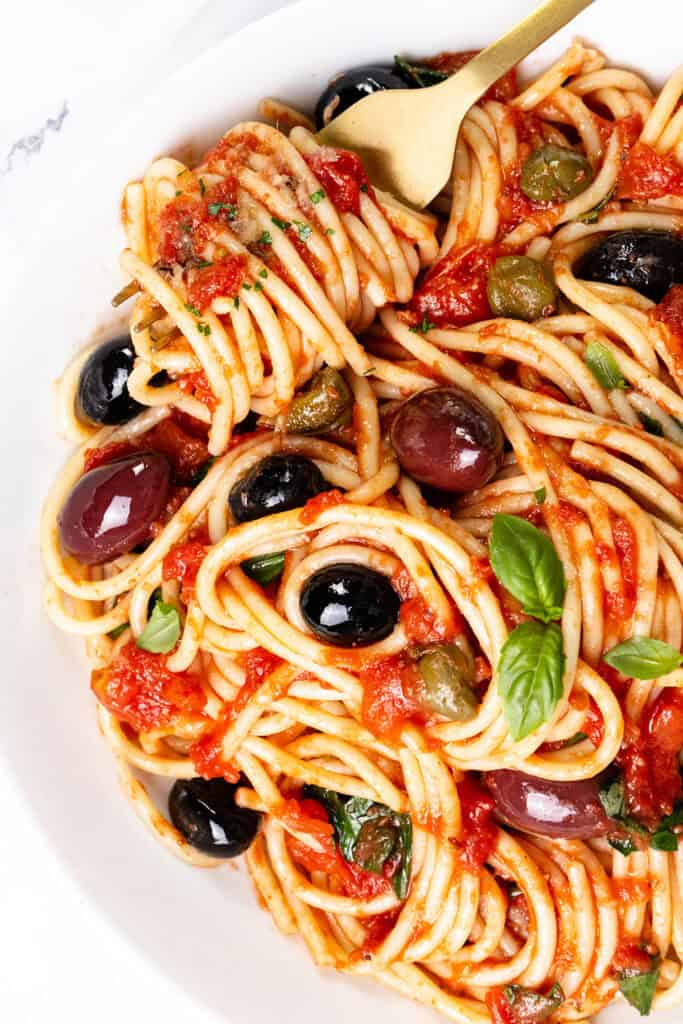 A bowl of Spaghetti Alla Puttanesca tossed in tomato sauce with black and green olives, capers, and fresh basil leaves. A fork twirls some pasta at the top.