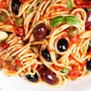 A close-up of Spaghetti Alla Puttanesca showcases a vibrant tomato sauce with black and green olives, capers, and fresh basil leaves. A gold fork elegantly twirls the pasta on a white plate, inviting you to indulge in its rich flavors.