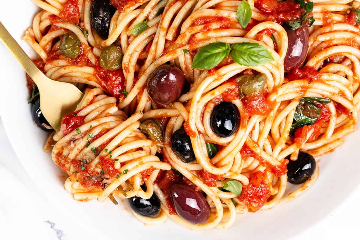 A close-up of Spaghetti Alla Puttanesca showcases a vibrant tomato sauce with black and green olives, capers, and fresh basil leaves. A gold fork elegantly twirls the pasta on a white plate, inviting you to indulge in its rich flavors.