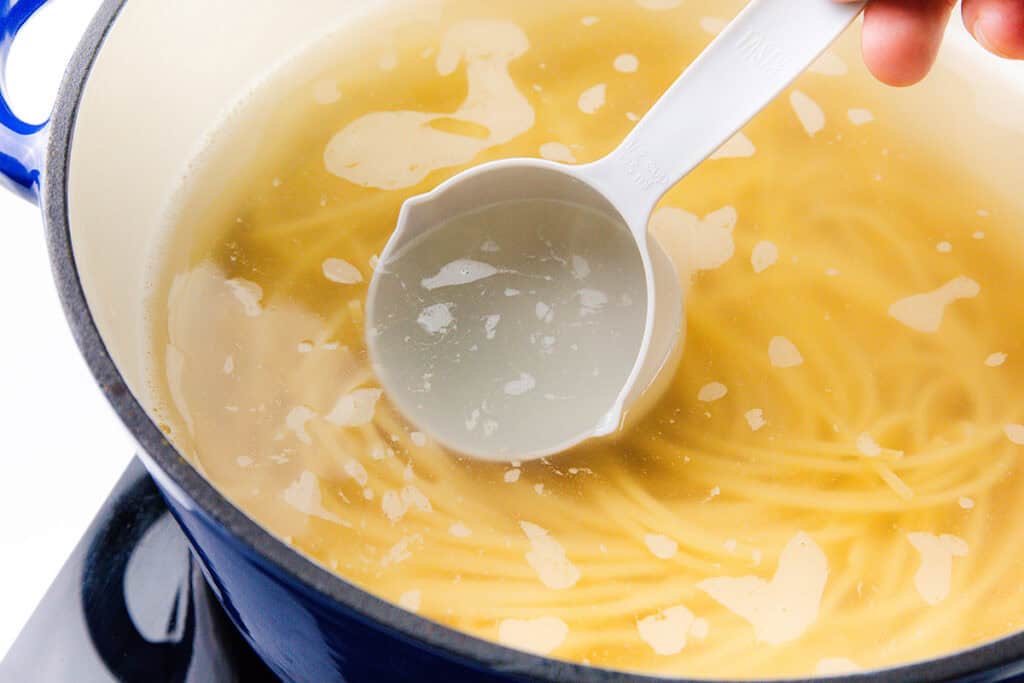 A blue pot filled with spaghetti cooking in boiling water, a nod to Spaghetti Alla Puttanesca. A hand holds a white ladle, scooping some starchy pasta water from the surface's swirling bubbles.