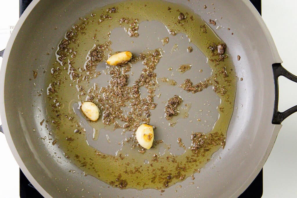 A frying pan with oil, garlic cloves, and brown spices sizzles on the stovetop, the aromas mingling as they sauté in preparation for a flavorful Spaghetti Alla Puttanesca.