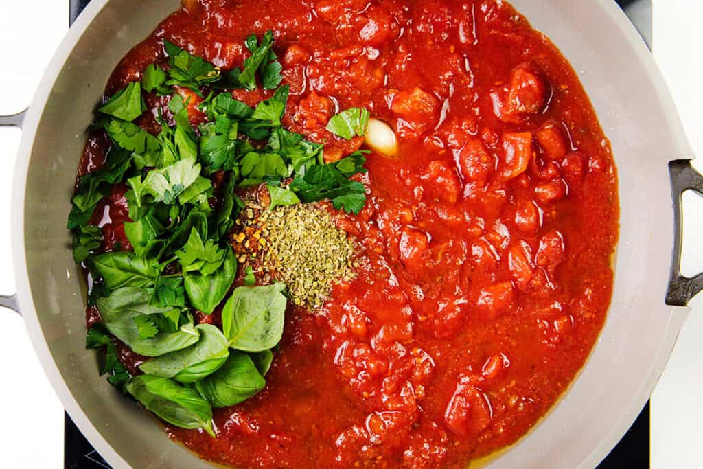A pan filled with chunky tomato sauce, fresh basil leaves, parsley, oregano, and garlic cloves evokes the start of Spaghetti Alla Puttanesca. The ingredients are neatly arranged with herbs on one side and sauce on the other, ready to be mixed and cooked.