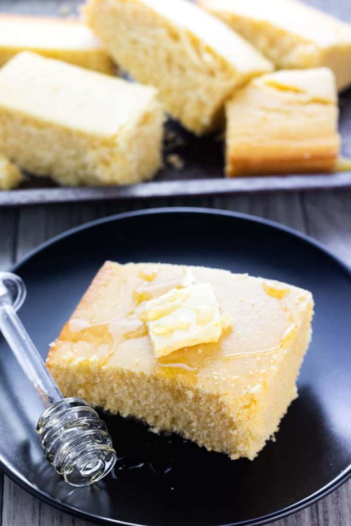 A piece of Cornbread on a plate topped with butter and drizzled with honey