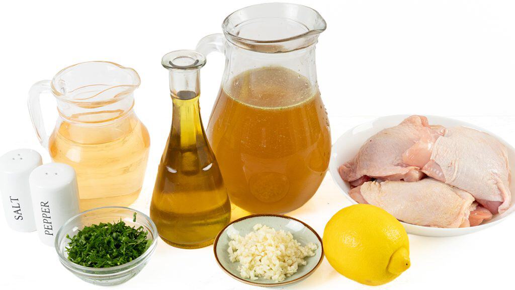 A bowl of raw chicken thighs, perfect for making Baked Garlic Chicken Thighs, sits alongside a lemon, chopped garlic, parsley, a small jug of broth, olive oil, white wine, and salt and pepper shakers arranged on a white background.