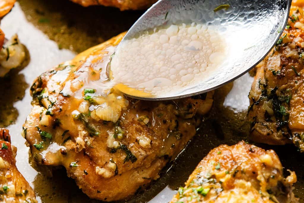 A spoon drizzles garlic herb butter sauce over a piece of cooked Baked Garlic Chicken Thighs, garnished with chopped herbs.