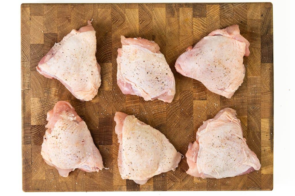 Six raw, seasoned chicken thighs arranged in two rows on a wooden cutting board, ready to be transformed into flavorful Baked Garlic Chicken Thighs.