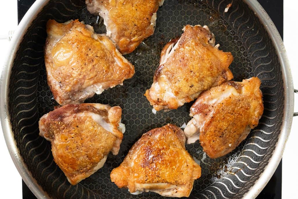 Six golden-brown Baked Garlic Chicken Thighs with crispy skin are sizzling in a nonstick frying pan on a stovetop.